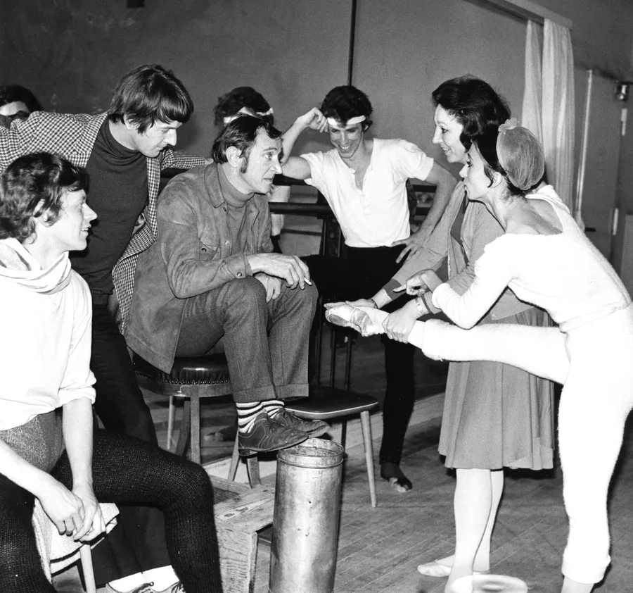 Schwarzweißfoto einer Ballettprobe: Choreograf John Cranko sitzt auf einem Stuhl, umgeben von Tänzerinnen und Tänzern. Eine Ballerina zeigt ihm ihre Fußposition, während die Gruppe aufmerksam und gut gelaunt zuhört.