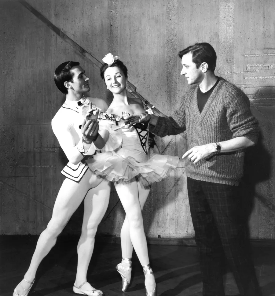 Schwarzweißfoto: Choreograf John Cranko gibt einem Tänzerpaar Anweisungen. Der Tänzer und die Ballerina tragen klassische Bühnenkostüme und stehen in einer eleganten Pose. Cranko trägt einen Pullover und Hosen und zeigt lächelnd mit der Hand eine Bewegung an.